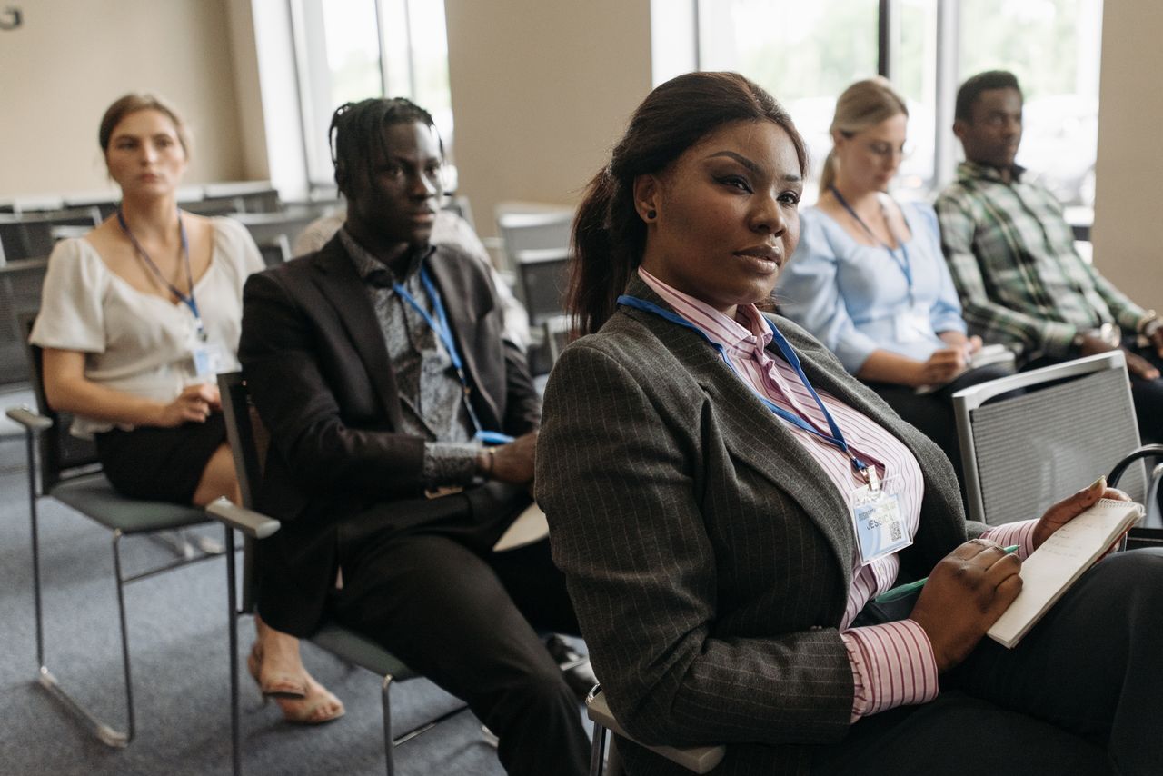 A diverse group attentively listens in a business seminar setting, indoors with notebooks.